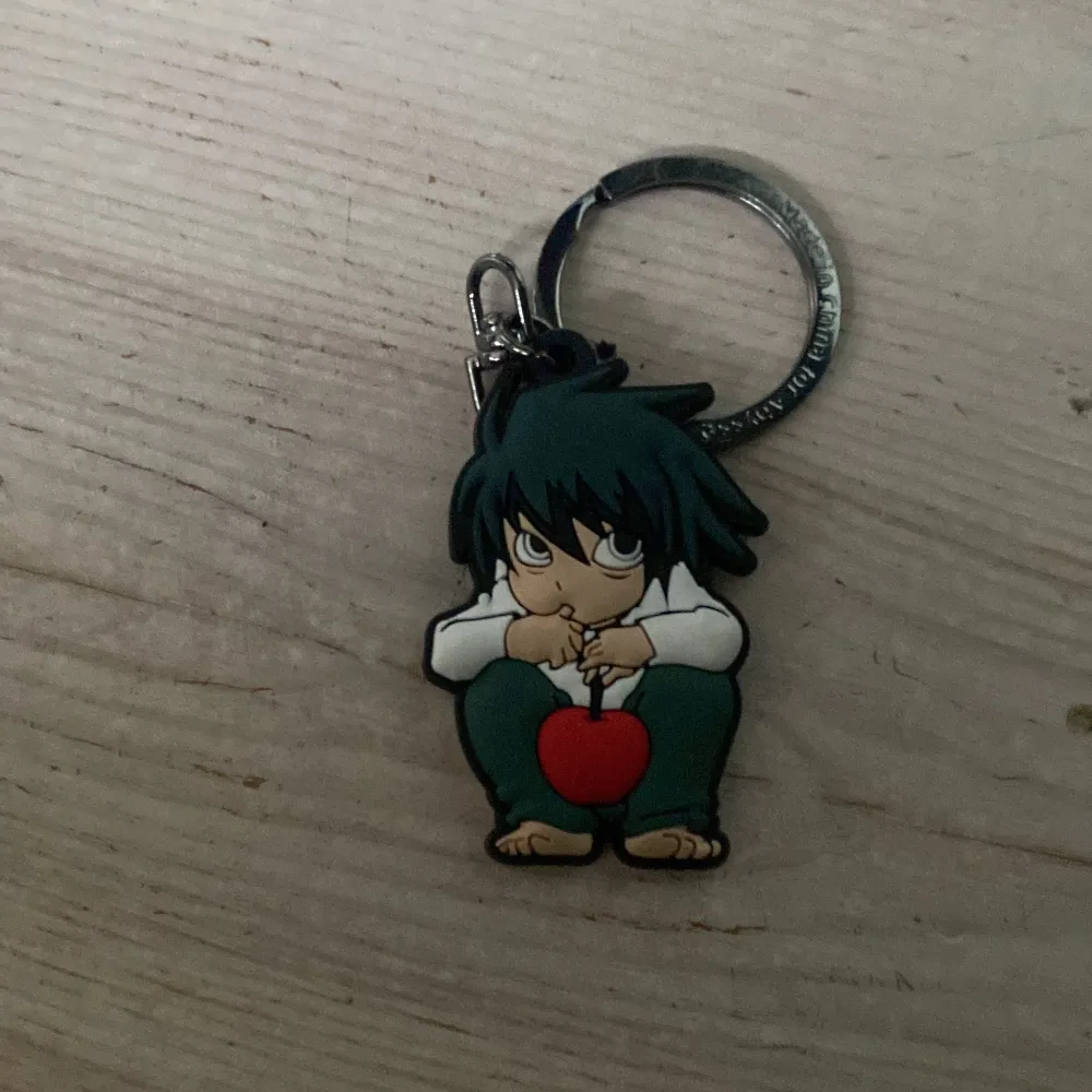 Cool nyckelring med en animefigur som sitter med ett rött äpple. Figuren har mörkt hår och bär en vit tröja och gröna byxor. Perfekt för fans av anime och manga.. Asusteet.