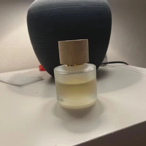 Parfym med trälock - Elegant parfymflaska med frostat glas och ett stilrent trälock. Flaskan har en rund form och innehåller en ljusgul vätska. Perfekt för den som uppskattar en minimalistisk design.