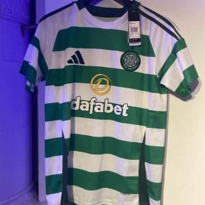 Grön och vit randig fotbollströja från Adidas - Säljer en grön och vit randig fotbollströja från Adidas med korta ärmar. Tröjan har en klubbemblem på bröstet och sponsorlogga på framsidan. Perfekt för fotbollsfans!