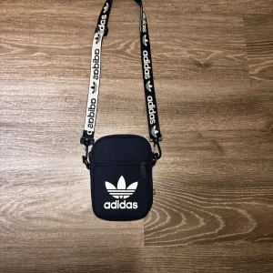 (Helt ny och oanvänd) axelväska från Adidas - Snygg svart axelväska från Adidas med justerbar rem och dragkedja. Perfekt för att bära med sig det nödvändigaste. Väskan har Adidas-loggan tryckt i vitt på framsidan och en rem med Adidas-mönster.