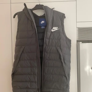 Grå dunväst från Nike - Säljer en grå dunväst från Nike. Västen har en dragkedja framtill och luva bak. Den är i storlek S och har en klassisk Nike-logga på bröstet. 