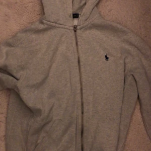 Grå hoodie från Polo Ralph Lauren - Säljer en snygg och bekväm grå zip hoodie från Polo Ralph Lauren. Perfekt för en avslappnad stil eller som ett extra lager under kalla dagar. OBS!!! saknar snöre och dragkedjan åker ner av sig själv ibland till över mitten. Inga fläckar!