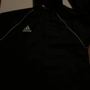 Svart träningsjacka från Adidas - Säljer en svart träningsjacka från Adidas i storlek M. Jackan har en dragkedja framtill och är långärmad. Perfekt för träning eller som en lätt jacka till vardags. Den är i bra skick och har de klassiska Adidas-ränderna längs ärmarna.