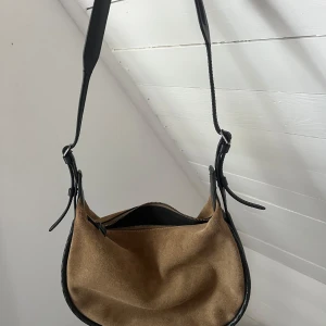 Beige och svart axelväska - Säljer en snygg beige axelväska med svarta detaljer. Väskan har en dragkedja upptill och är perfekt för vardagsbruk. Den är i bra skick och har en justerbar axelrem för bekväm passform. Perfekt för att bära dina dagliga nödvändigheter!