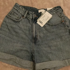 Blå jeansshorts från H&M - Säljer ett par snygga blå jeansshorts från H&M. De är högmidjade med en klassisk mom fit och har uppvikta ben. Perfekta för sommardagar och kan lätt matchas med en t-shirt eller linne. De har fickor både fram och bak, och stängs med dragkedja och knapp.
