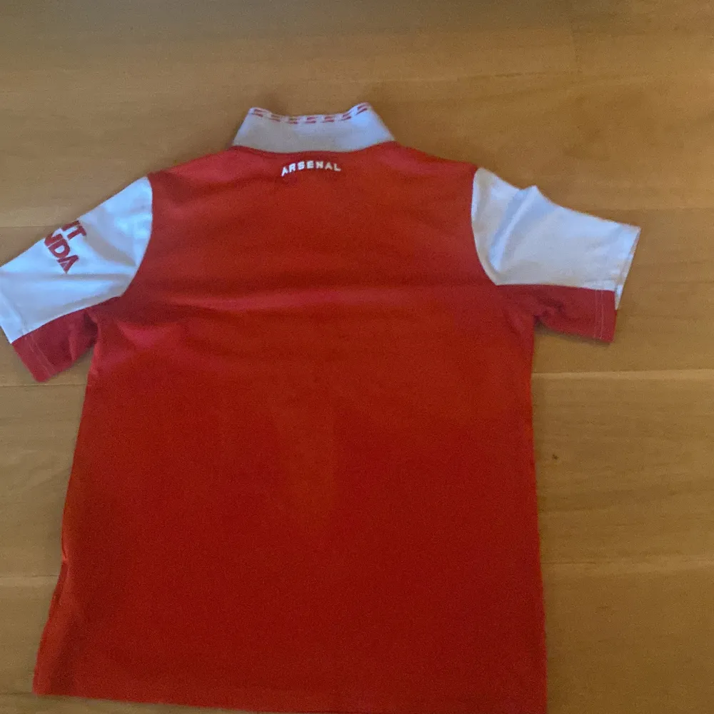 Säljer en snygg röd Arsenal fotbollströja från Adidas. Den har korta vita ärmar och det klassiska Arsenal-emblemet på bröstet. Tröjan är i storlek 164, vilket passar perfekt för ungdomar. Den har även sponsorloggan 'Emirates Fly Better' tryckt på framsidan. Perfekt för alla Arsenal-fans!. T-shirts.