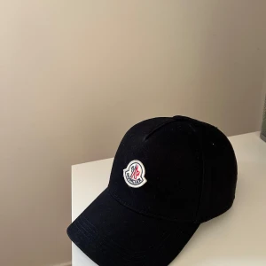 Svart keps från Moncler - Säljer en svart keps. Den har en broderad logga framtill och justerbar rem baktill för perfekt passform. Kepsen är i bra skick och passar perfekt för vardagen. Perfekt accessoar för att lyfta din stil! Kvitto finns ej. Pris kan diskuteras.