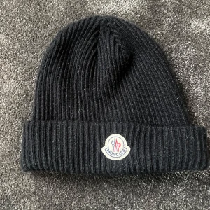 Svart ribbad mössa från Moncler - Säljer en snygg svart mössa från Moncler. Den är perfekt för kalla dagar och har en stilren design med Monclers logga framtill. Mössan är i bra skick och passar till de flesta outfits. Perfekt för vintern! ❄️