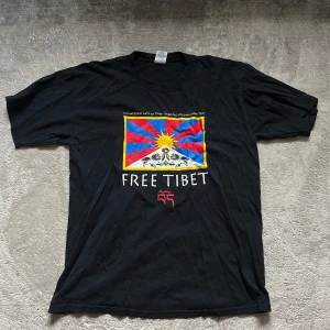 Svart T-shirt med Free Tibet-tryck - Säljer en svart T-shirt med ett stort färgglatt tryck av Tibet-flaggan och texten 'FREE TIBET' under. T-shirten är i bomull och har en normal passform med korta ärmar. Perfekt för att visa ditt stöd för Tibet!