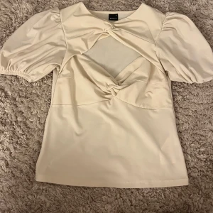 Beige topp från Gina Tricot - Säljer en superfin beige topp från Gina Tricot i storlek M. Den har korta puffärmar och en snygg knytdetalj framtill som ger en unik look. Perfekt för både vardag och fest!