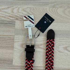 Flätat bälte från Ben Sherman - Säljer ett snyggt flätat bälte från Ben Sherman i rött, svart och vitt. Bältet har en klassisk design med ett silverfärgat spänne och bruna skinnändar. Perfekt för att ge en färgklick till din outfit!