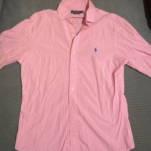 Rosa skjorta från Ralph Lauren - Snygg rosa skjorta från Ralph Lauren i slim fit. Storlek M. Tillverkad i 100% bomull med klassisk krage och knappar framtill. Den har en broderad blå logotyp på bröstet som ger en stilren touch. Perfekt för en stilmedveten look!