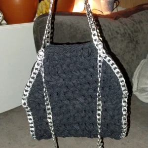 Svart virkad väska med kedjedetaljer - Snygg svart virkad väska med silverfärgade kedjedetaljer. Perfekt för att ge din outfit en unik touch. Väskan har en enkel design med två kedjehandtag som ger en cool kontrast till det mjuka materialet.
