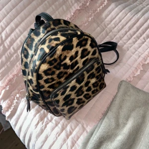Leopardmönstrad ryggsäck med dragkedja - Snygg leopardmönstrad ryggsäck med flera dragkedjor och justerbara axelremmar. Perfekt för att ge din outfit en vild touch. Ryggsäcken har en praktisk framficka och är tillverkad i syntetmaterial.