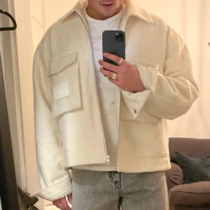 Calvin Klein Jacka - Säljer en stilren beige fodrad overahirt med dragkedja och stora fickor framtill. Jackan har en klassisk krage och långa ärmar med knappar vid ärmsluten. Perfekt för lager-på-lager-stil under höst och vår.