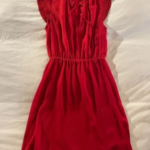 Röd klänning från H&M - Snygg röd klänning från H&M med korta ärmar och knytning vid halsen. Den har en elastisk midja för en bekväm passform och är perfekt för en färgstark look. Klänningen är gjord i ett lätt och luftigt material.