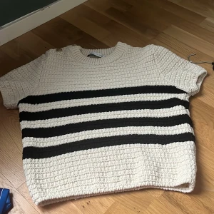 Randig stickad tröja - Säljer en mysig stickad tröja med ett klassiskt randigt mönster i svart och beige. Tröjan har korta ärmar och en rund halsringning. Perfekt för en avslappnad stil.