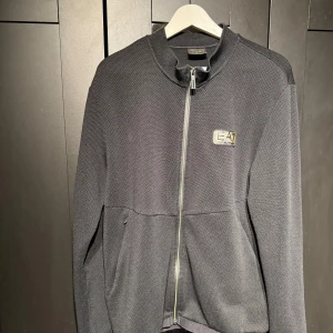 EA7 tröja  - Snygg svart tröja från EA7 med dragkedja och broderad logga på bröstet. Tröjan har en stilren design med långärmade ärmar och en bekväm passform. Perfekt för en casual look.