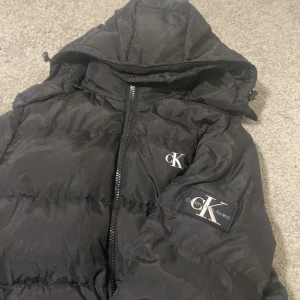 Svart pufferjacka från Calvin Klein Jeans - Storlek xs passar 169 cmSäljer en snygg svart pufferjacka från Calvin Klein Jeans. Jackan har en dragkedja framtill och en huva för extra skydd. Den är dekorerad med CK-loggan på bröstet och ärmen. Perfekt för kyliga dagar!