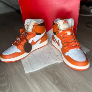 Wmns Air Jordans 1 Retro HI OG  orange och vit - Snygga Nike Air Jordans i en fräsch kombination av orange och vit. Skorna har hög snörning och en klassisk design med Nike-loggan på sidan. Perfekta för dig som vill sticka ut med stil.