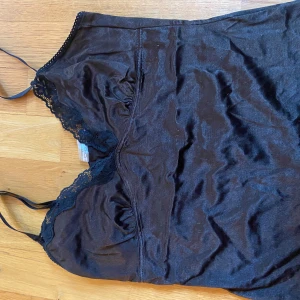 Svart linne från Pieces - Säljer ett svart linne från Pieces med spetsdetaljer vid halsringningen. Linnet har tunna axelband och en mjuk, glansig yta. Perfekt för en elegant look.