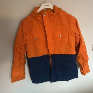 Orange och marinblå jacka från Burton - Säljer en cool orange och marinblå jacka från Burton. Jackan har en huva och praktiska fickor med knappar framtill. Perfekt för utomhusaktiviteter med sin funktionella design och snygga färgkombination.