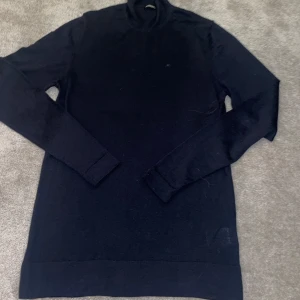 Svart merinoull polotröja från J.Lindeberg - Säljer en stilren svart polotröja från J.Lindeberg i 100% merinoull. Tröjan har en klassisk design med lång ärm och en liten broderad logotyp på bröstet. Perfekt för kyligare dagar.