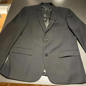 Svart kavaj från Dressmann - Snygg svart kavaj från Dressmann i slim fit. Tillverkad av 65% polyester och 35% viskos med ett foder av 100% polyester. Perfekt för en stilren look med två knappar framtill och klassiska fickor. Passar utmärkt för både formella och semi-formella tillfällen.