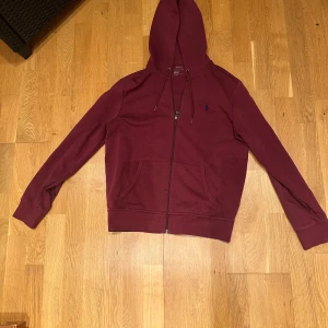 Vinröd hoodie från Ralph Lauren - Säljer en snygg vinröd hoodie från Ralph Lauren med dragkedja och justerbar huva. Den har en liten broderad logga på bröstet och är tillverkad i ett mjukt material. Perfekt för en avslappnad stil.