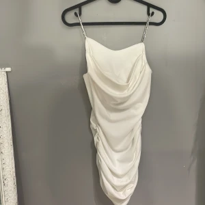 Vit klänning med glitter band - Elegant vit axelbandslös klänning med draperad design och smala axelband. Perfekt för en stilren look med en touch av glamour. Klänningen har en åtsittande passform som framhäver figuren.