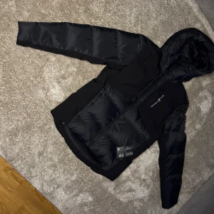 Svart dunjacka från Sail Racing - Säljer en svart dunjacka från Sail Racing med 500 fill power. Jackan har en dragkedja framtill och en bröstficka med dragkedja. Perfekt för kyliga dagar med sin quiltade design och långa ärmar.