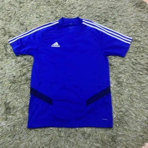 Blå fotbollströja från Adidas - Snygg blå fotbollströja från Adidas med klassiska vita ränder på axlarna. Tröjan är kortärmad och tillverkad i ClimaCool-material för optimal ventilation. Perfekt för träning eller match!