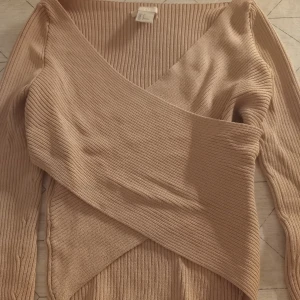 Beige ribbad omlottopp från H&M - Snygg beige ribbad omlottopp från H&M i storlek XS. Toppen har en unik omlottdesign och långa ärmar, perfekt för en stilren look. Passar bra till både jeans och kjol.