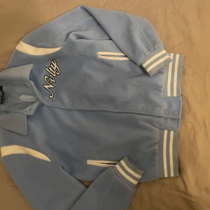 Ljusblå fleecejacka från NVLTY - Snygg ljusblå fleecejacka från NVLTY med vita detaljer och texten 'Nvlty' broderad på både fram- och baksidan. Jackan har långa ärmar och ribbade muddar samt knappar framtill. Perfekt för en cool och avslappnad stil.