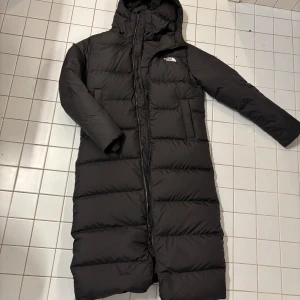 Svart dunjacka från The North Face - Säljer en lång svart dunjacka från The North Face. Jackan har en huva och dragkedja framtill. Perfekt för kalla dagar med sin vadderade design som håller dig varm och bekväm.Helt ny inköpt förra veckan kvitton finns nypris 6000 stl S