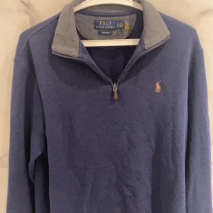 Blå quarter-zip tröja från Polo Ralph Lauren - Säljer en stilren blå quarter-zip tröja från Polo Ralph Lauren. Tröjan har en broderad logotyp på bröstet och en kontrasterande grå krage. Perfekt för en avslappnad och snygg look. Skicket är på 8/10 nypris 2195kr