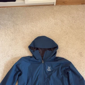 Blå vindjacka från Haglöfs - Säljer en blå vindjacka från Haglöfs med huva och dragkedja. Perfekt för utomhusaktiviteter med sin funktionella design. Jackan har en stilren look och är tillverkad i slitstarkt material.