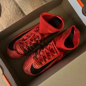 Röda Nike Mercurial fotbollsskor - Snygga röda Nike Mercurial fotbollsskor med svart swoosh och snörning. Perfekta för fotbollsplanen med sina greppvänliga dobbar och strumpliknande passform.