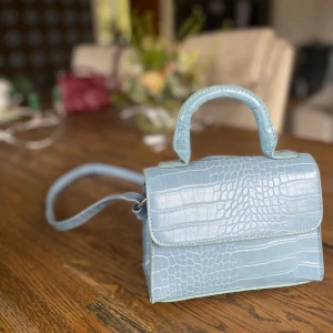 Ljusblå axelväska med krokodilmönster - Snygg ljusblå axelväska med krokodilmönster. Väskan har ett praktiskt handtag och en justerbar axelrem.