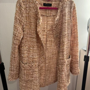 Beige kappa från Risskio - Säljer en elegant beige kappa från Risskio. Den har en texturerad yta med en öppen design och två fickor framtill. Perfekt för en stilren look. Knappt använd!