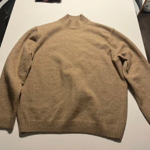 Beige stickad tröja från Weekday - Säljer en stilren beige stickad tröja från Weekday. Tröjan har en hög ribbad krage och långa ärmar, perfekt för kyligare dagar. Den är mjuk och bekväm, idealisk för både vardag och avslappnade tillfällen.