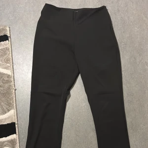 Svarta byxor från Gina Tricot - Snygga svarta byxor från Gina Tricot med dragkedja i sidan. Perfekta för en stilren look. Byxorna har en rak passform och är tillverkade i ett bekvämt material. Dragkedjan funkar men grejen man drar i är sönder