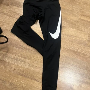 Svarta leggings från Nike - Snygga svarta leggings från Nike med Dri-FIT-teknologi. De har en stor vit Nike-logga längs benet och är perfekta för träning. Materialet är syntetiskt och designat för att hålla dig torr och bekväm.
