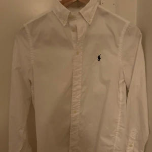 Vit skjorta från Ralph Lauren - Klassisk vit skjorta från Ralph Lauren med broderad logga på bröstet.  Skjortan är i storlek 14-16 och passar någon som är 160-165.Använd ett fåtal gånger och är i bra skick. Nypris: 1300kr. Tveka inte att höra av sig vid minsta fundering.