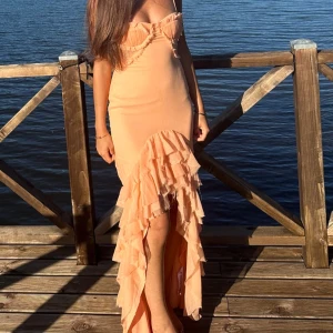 Persikofärgad klänning med volanger - Säljer en elegant persikofärgad klänning som används 1 gång med smala axelband och volanger framtill. Klänningen har en lång design med en slits som ger en fin rörelse. Perfekt för somriga tillfällen vid vattnet. 🌸