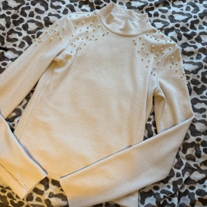 Beige  ribbad polotröja med pärlor - Säljer en beige ribbad polotröja dekorerad med pärlor på axlarna. Tröjan har långa ärmar och en hög krage som ger en elegant look. Perfekt för kyligare dagar. (Aldrig använt)