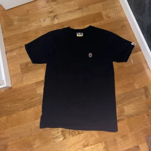 Svart t-shirt från A Bathing Ape - Säljer en svart t-shirt från A Bathing Ape med ett litet brunt och vitt logomärke på bröstet. T-shirten har en klassisk rund hals och korta ärmar. Perfekt för en stilren och avslappnad look.