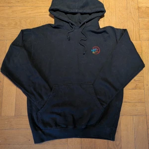 Svart GRIP skate hoodie - Säljer en svart hoodie från skatemärket GRIP med en cool broderad logga i flera färger på bröstet och färgglada ord på ryggen. Perfekt för dig som vill ha en unik och stilren look. Hoodien har en klassisk passform med känguruficka och justerbar huva. M/L Skick: Har bara använts några gånger, dammet och håret på bilderna kan tas bort enkelt