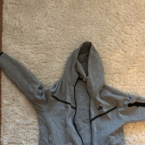 Grå hoodie och byxor från Nike - Säljer en grå hoodie och matchande byxor från Nike. Hoodien har dragkedja och svarta detaljer samt en luva. Perfekt för en avslappnad stil.