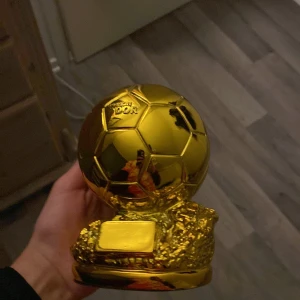 Guld Ballon d'Or trofé - En liten replika av den ikoniska Ballon d'Or trofén i guld. Perfekt för fotbollsälskare som vill ha en bit av fotbollshistorien hemma. Trofén har en detaljerad design med en fotboll på en stenliknande bas.